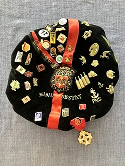 Une faluche vue de dessus : béret de velours noir, ruban rouge et ruban doré, écusson de ville brodé, devise, et des dizaines de pin's accrochés